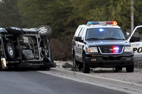 A Tucson Police patrol vehicle is parked just off the road facing the camera with the driver's side door open. Next to it, in the road, a passenger vehicle stands on its side, its front wheels twisted flat against the undercarriage.
