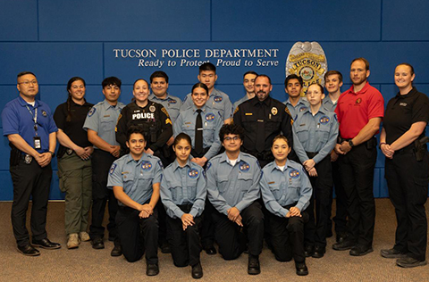 Tucson Explorers in uniform kneel and stand for a group photo with their advisors at TPD headquarters. The words Tucson Police Department, Ready to Protect, Proud to Serve, and a badge are on a blue wall behind them.