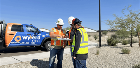 Three construction workers in safety gear review plans beside a Wyyerd Fiber truck in a desert-like area..
