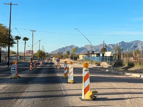 A sunlit street under construction with a line of orange-and-white traffic barriers and “Keep Right” signs dividing the road. Utility poles and palm trees line the sides, with small buildings and fenced lots nearby. Mountains rise in the background beneath a clear blue sky.