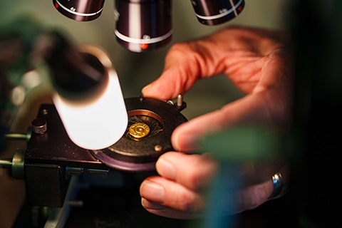 A man's hand positions a bullet in a NIBIN device under microscope lenses, a bright light in the foreground.