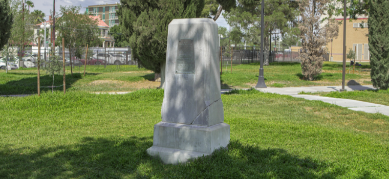 A weathered, blank stone monument stands in a grassy park. Bare trees and a building are in the background, conveying a quiet and solemn atmosphere.